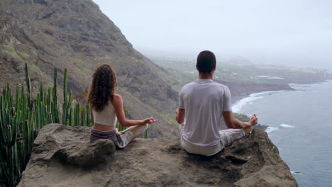 High-above-on-a-mountain,-a-man-and-a-woman-sit-on-stones,-meditating-with-lifted-hands,-taking-in-the-ocean-view,-and-practicing-soothing-breaths